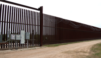 Border security fence along a dirt road with electrical equipment visible near the fence, set in an open landscape.
