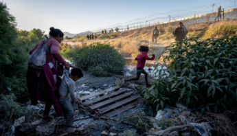 A mother helps her children cross a polluted area while migrants walk along a border fence in the background.